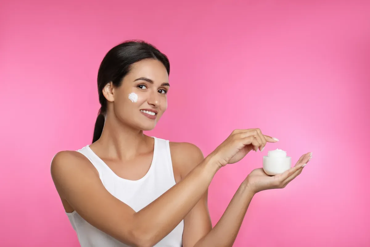 A smiling woman in a white vest holding a small jar of white cream, with a dab of cream on her cheek, against a pink background