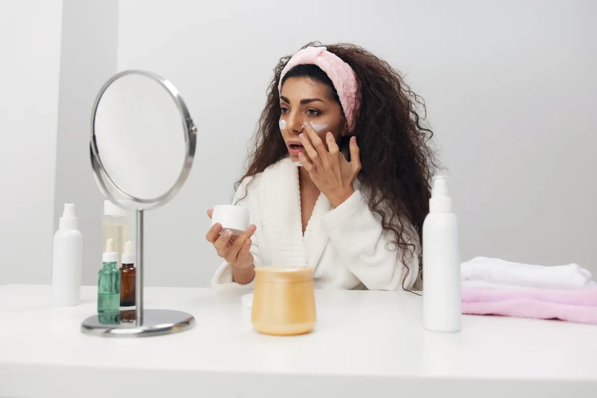 A woman in a white bathrobe and pink headband applying cream to her cheeks at a table with a round mirror, a cream jar and dropper bottles