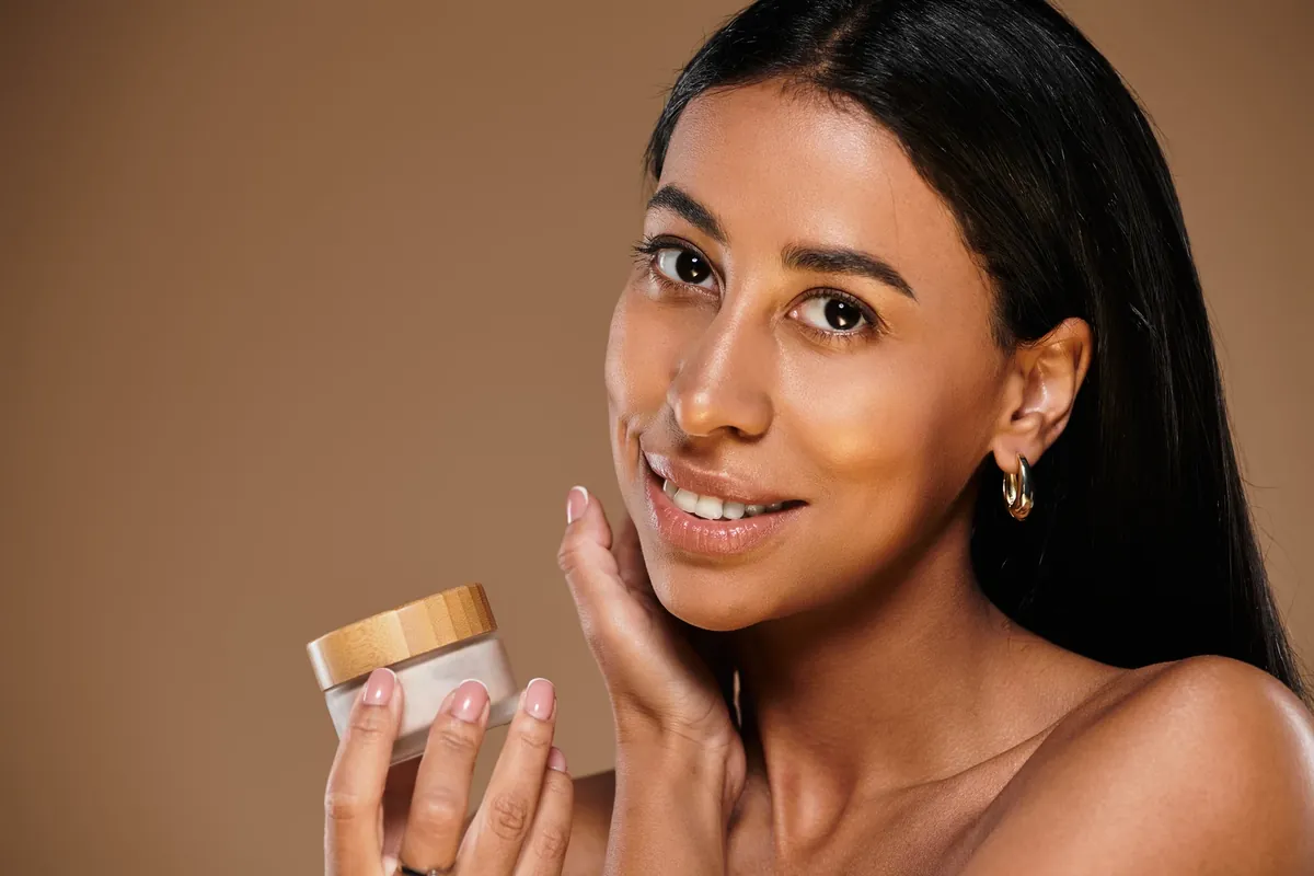 A smiling woman holds a small jar of cream with a wooden lid and touches her cheek against a brown backdrop