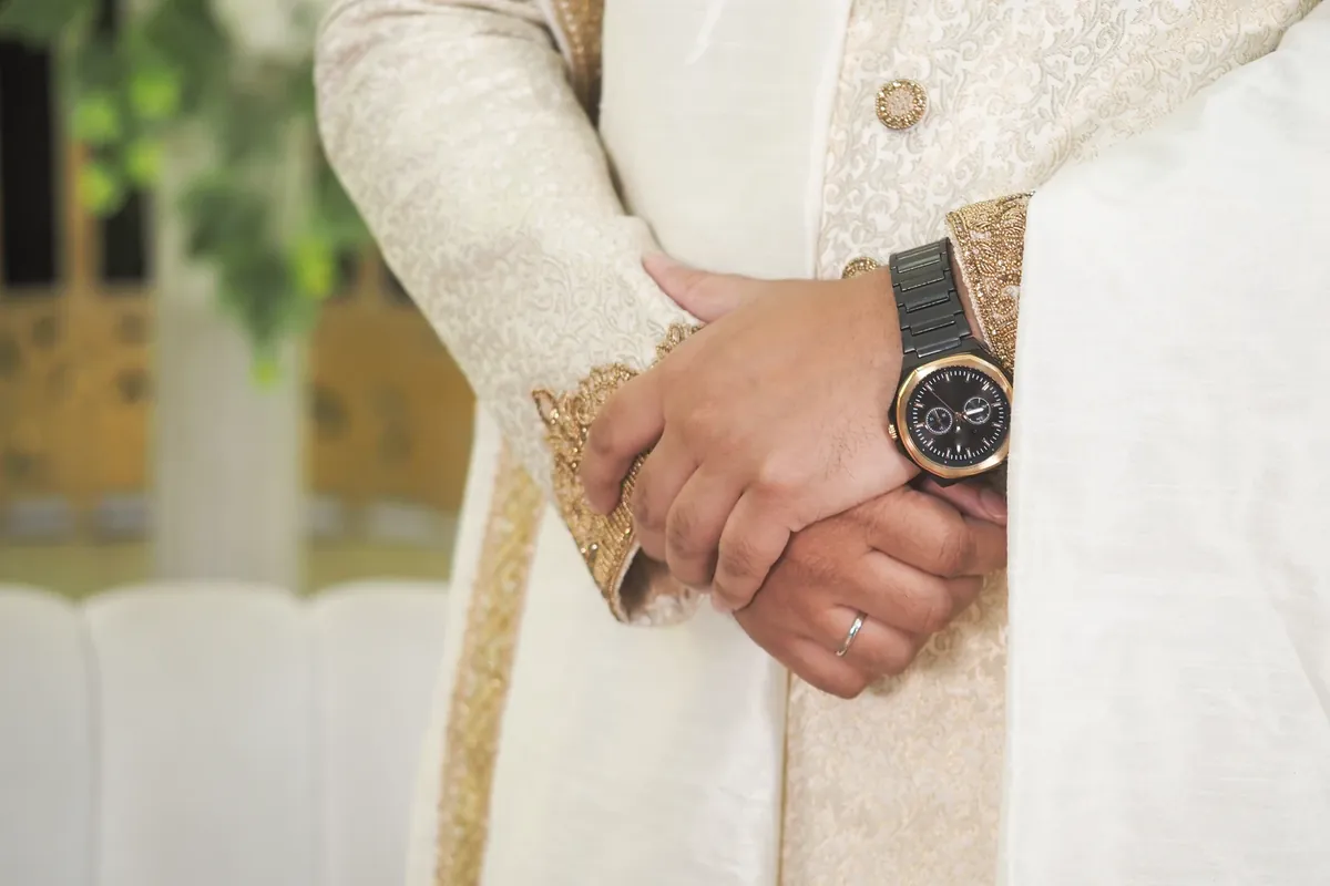 A man's hands clasped over the front of a cream embroidered sherwani, with a dark watch with a gold bezel on his wrist