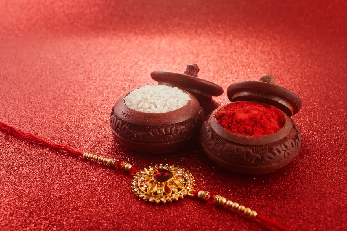 Two small carved wooden bowls, one filled with white rice and one with red powder, beside a red thread bracelet with a gold and red centrepiece, on a glittering red surface