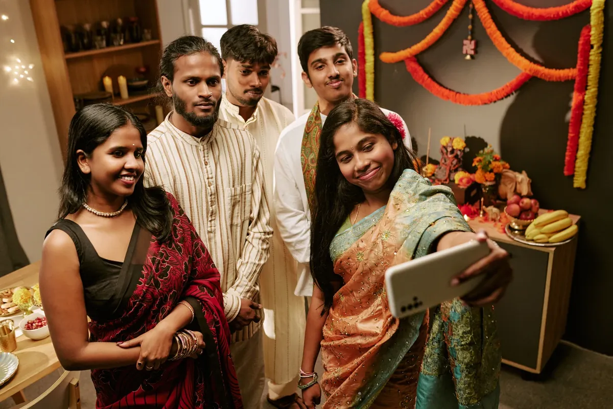 Five people in festive clothes crowd together to take a selfie in a room decorated with marigold garlands, with flowers and fruit on a table behind them