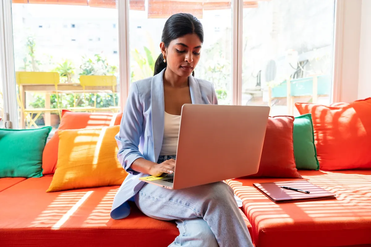A woman using a laptop on an orange sofa beside a bright window