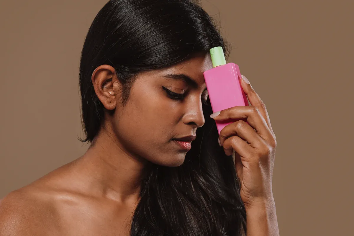 A woman holding a pink skincare bottle against her cheek