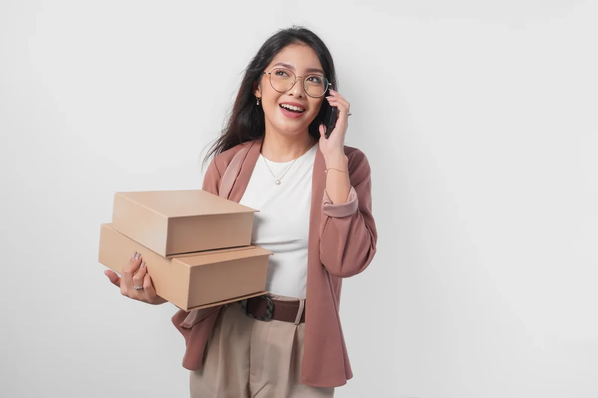 A smiling woman wearing glasses on a phone call, holding two cardboard parcels