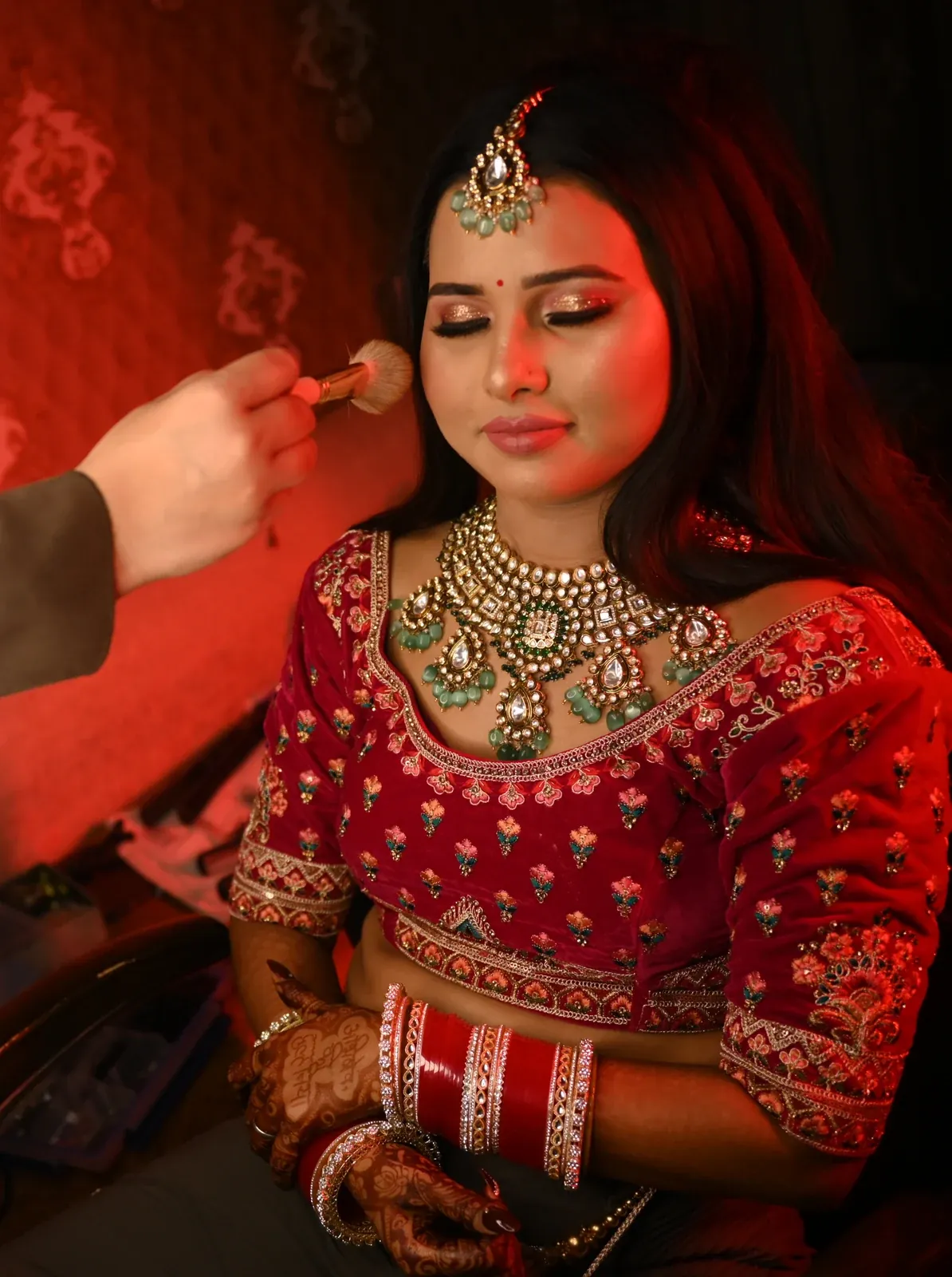 A bride in a red embroidered blouse wearing a kundan-style necklace with green drops and a maang tikka while a make-up artist's brush touches her cheek
