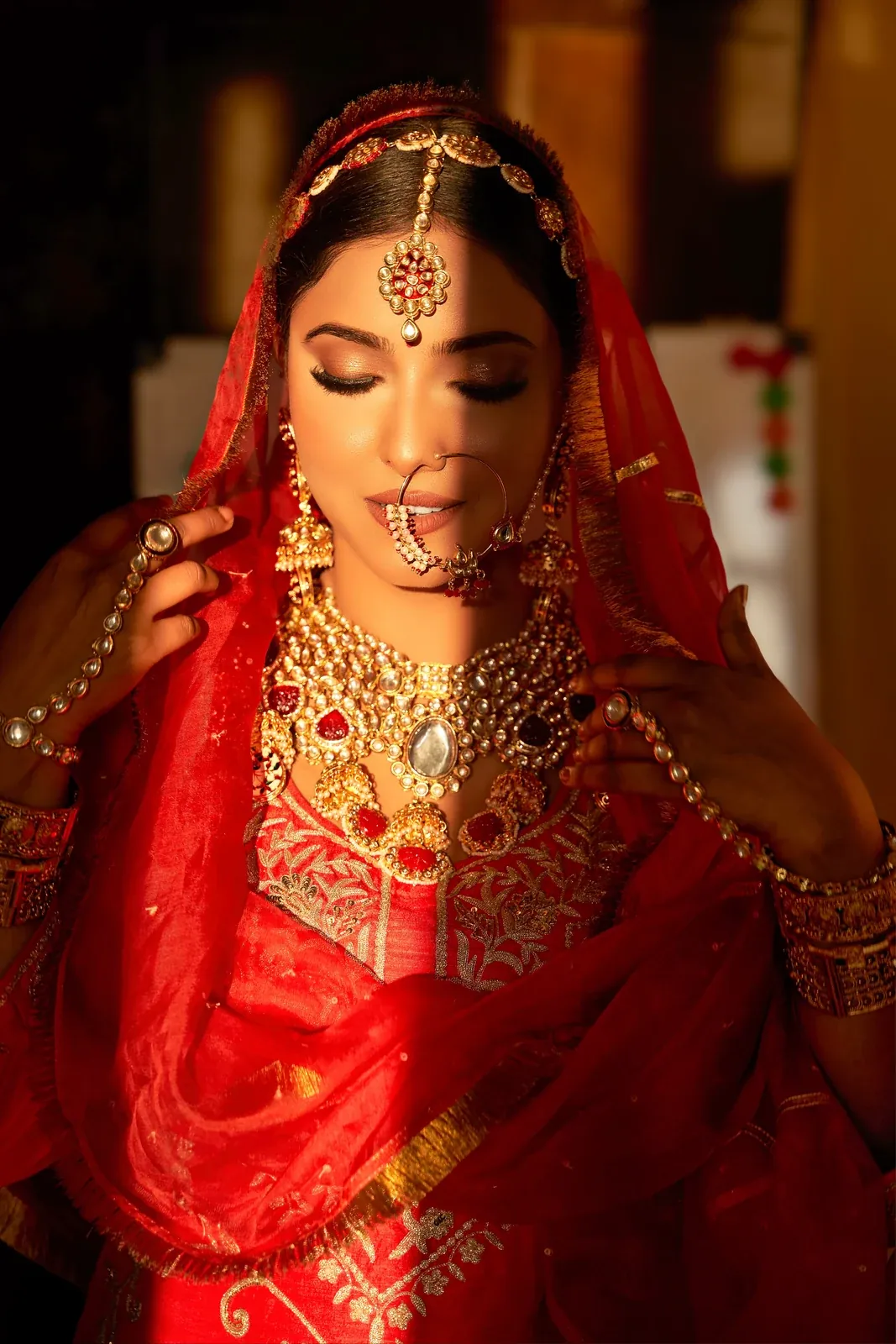 A bride in a red veil and outfit wearing a polki necklace, nose ring, maang tikka and jhumkas, eyes lowered