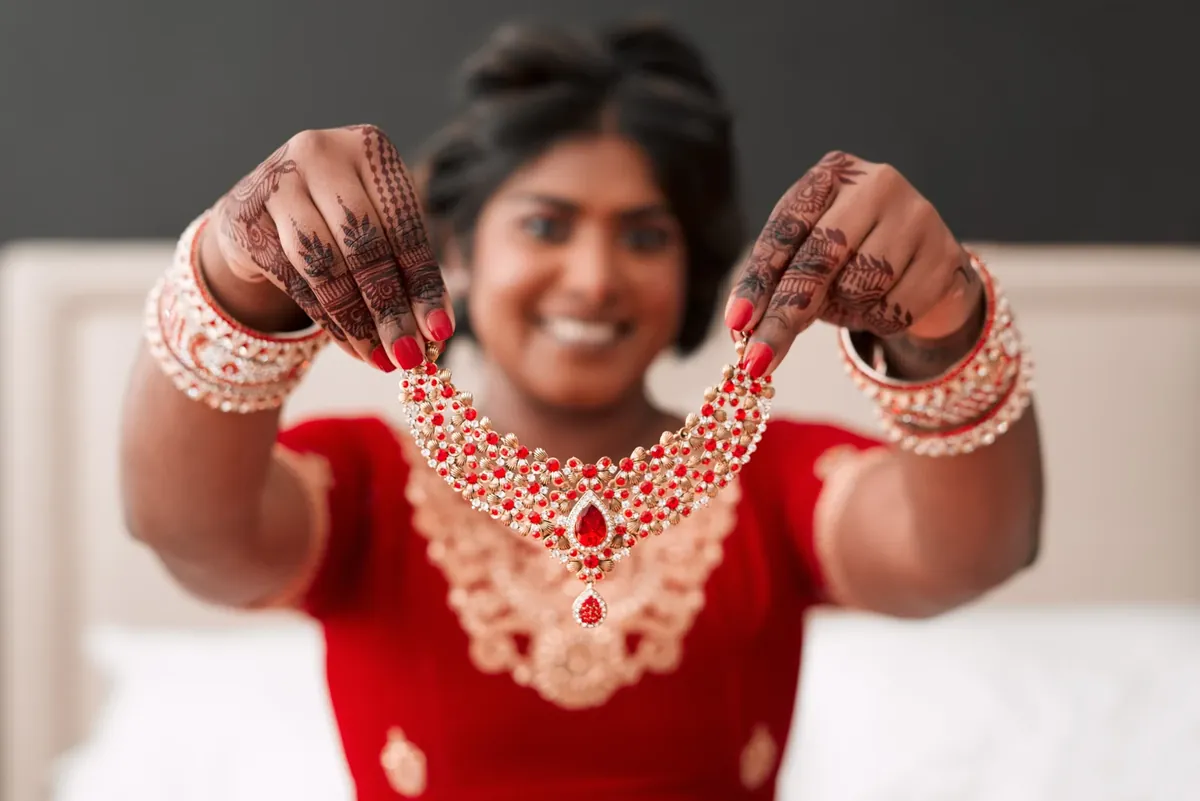 A smiling bride in red with henna on her hands holds up a red-stone and gold-bead bridal necklace
