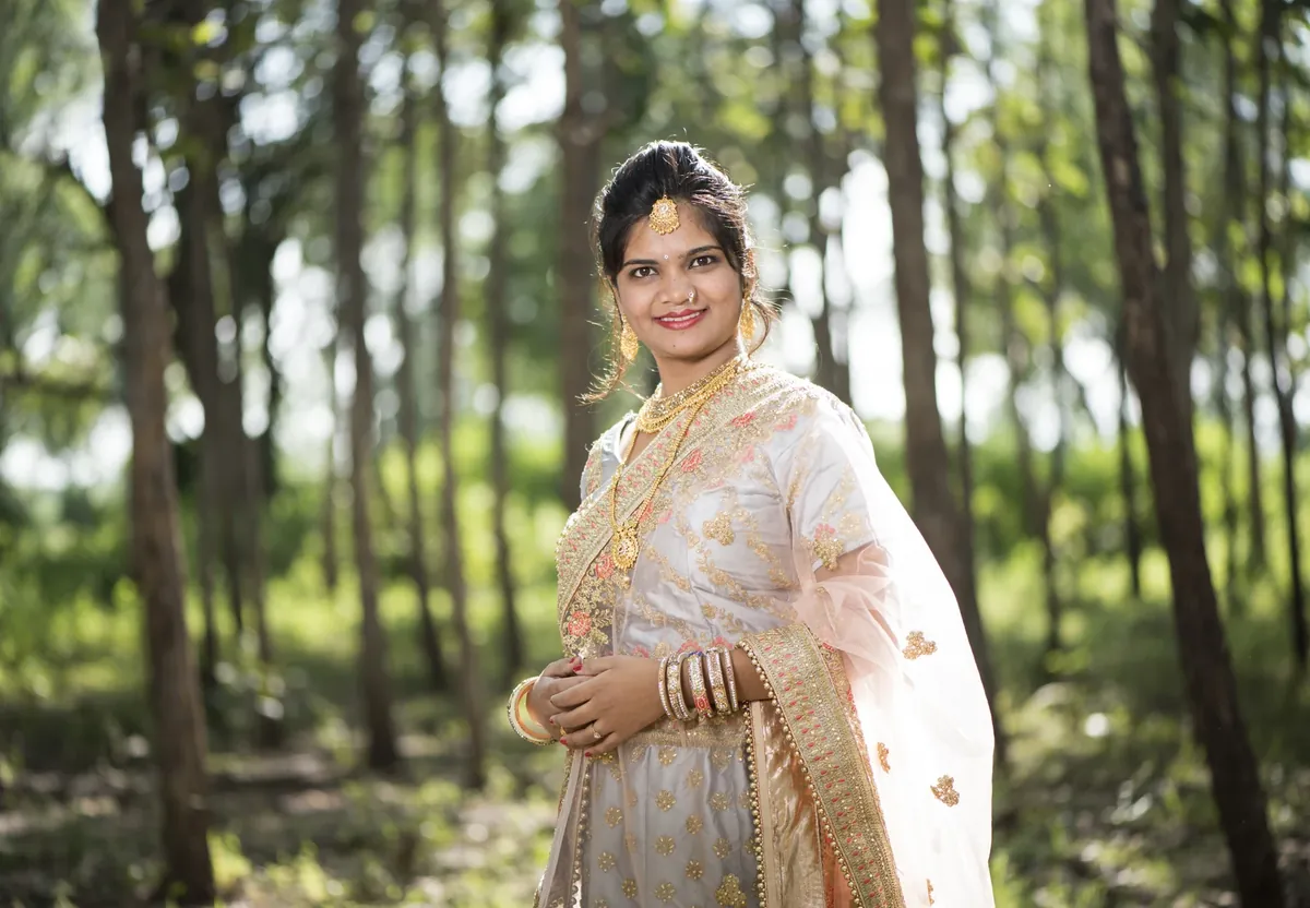 A woman in a cream lehenga with pink and gold embroidery, a net dupatta, gold jewellery and glass bangles, standing among tall trees