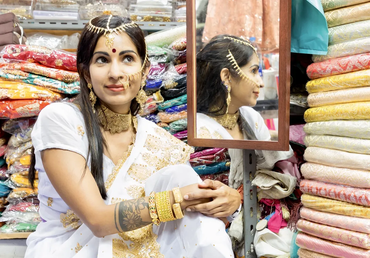 A woman in a white and gold outfit with gold jewellery sits beside a mirror in a fabric shop with stacked rolls of cloth