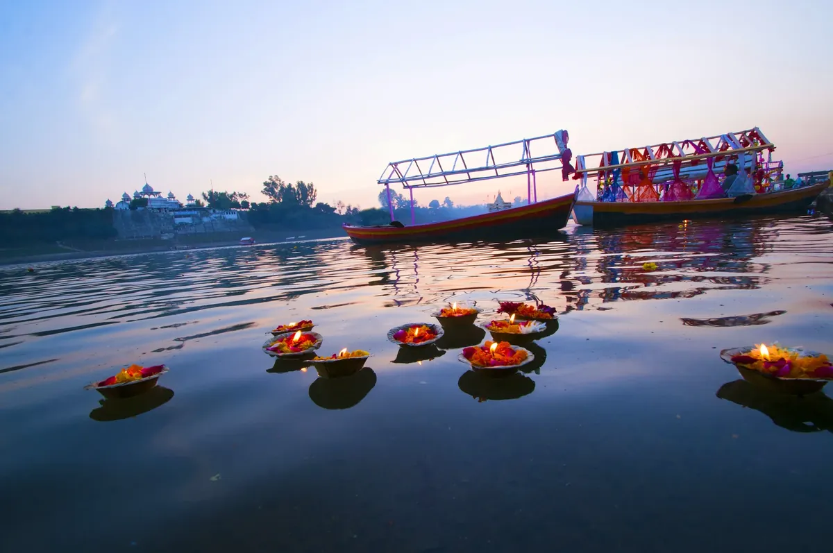 Small bowls of marigolds and lit lamps floating on calm river water at dusk, with two decorated boats behind