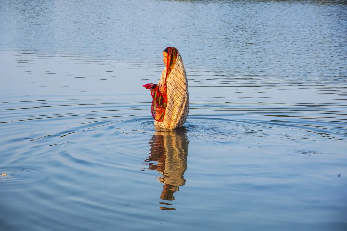 A woman wrapped in a checked shawl standing waist-deep in calm lake water, with her reflection beneath her