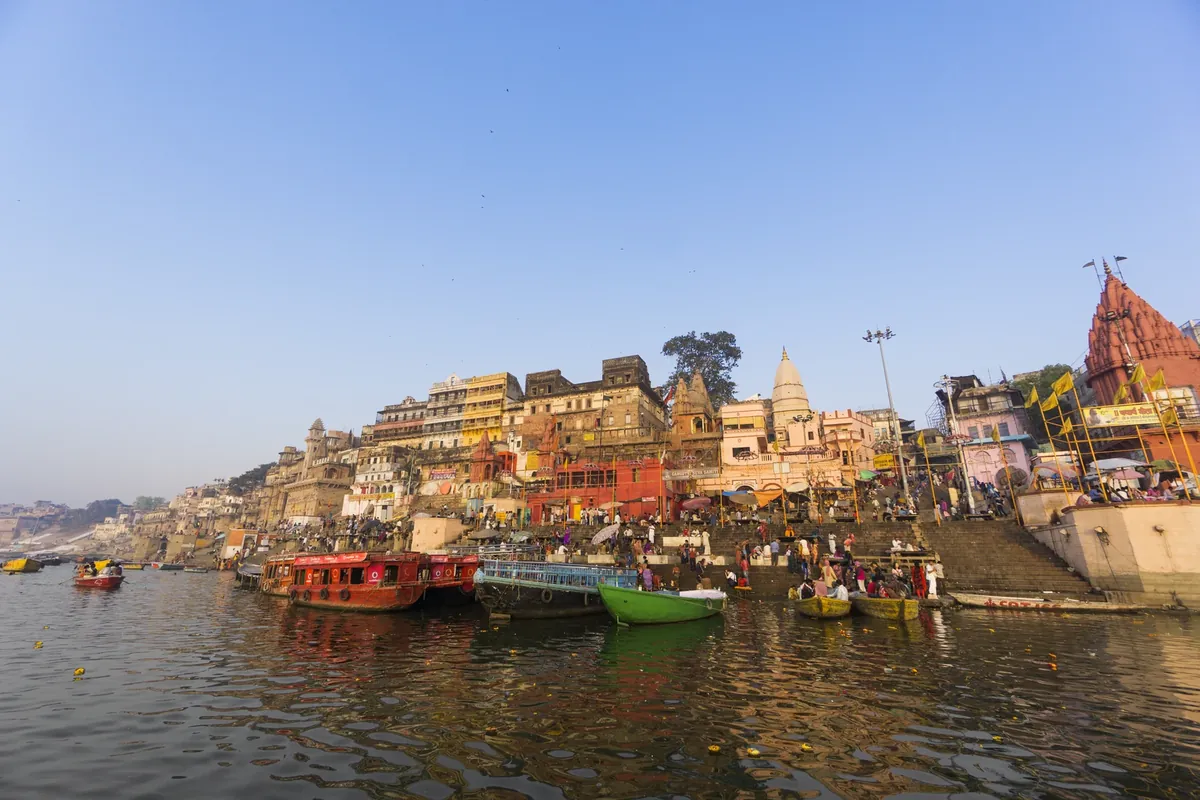 A view of the ghats at Varanasi from the river, with boats on the water and old buildings along the bank under a clear sky