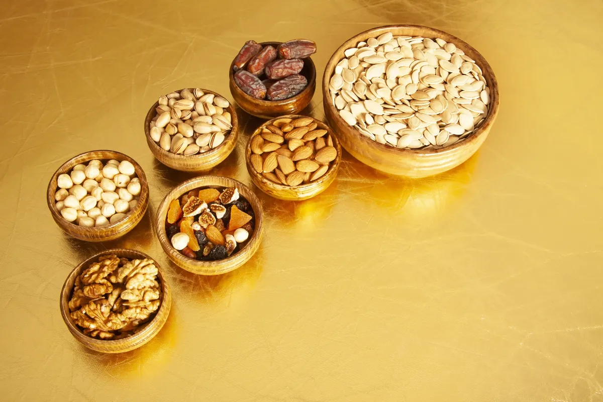 Wooden bowls of almonds, walnuts, pistachios, pumpkin seeds, dates and mixed dried fruit on a gold-coloured surface