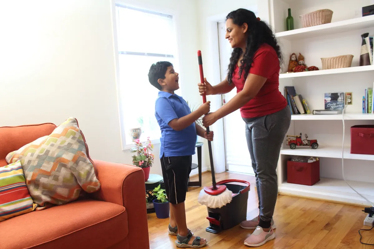A woman in a bright living room joking with a young boy while holding a mop and bucket