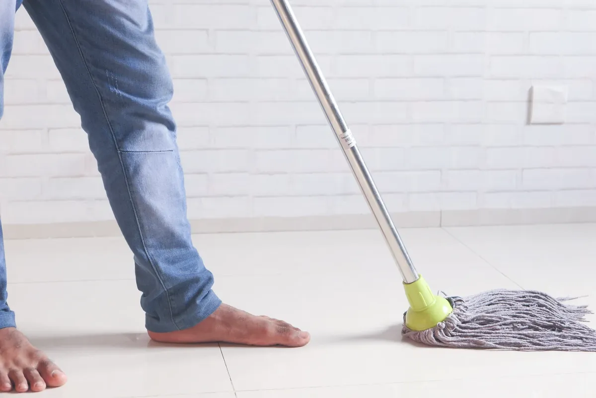 A person with bare feet mopping a tiled floor against a white brick wall