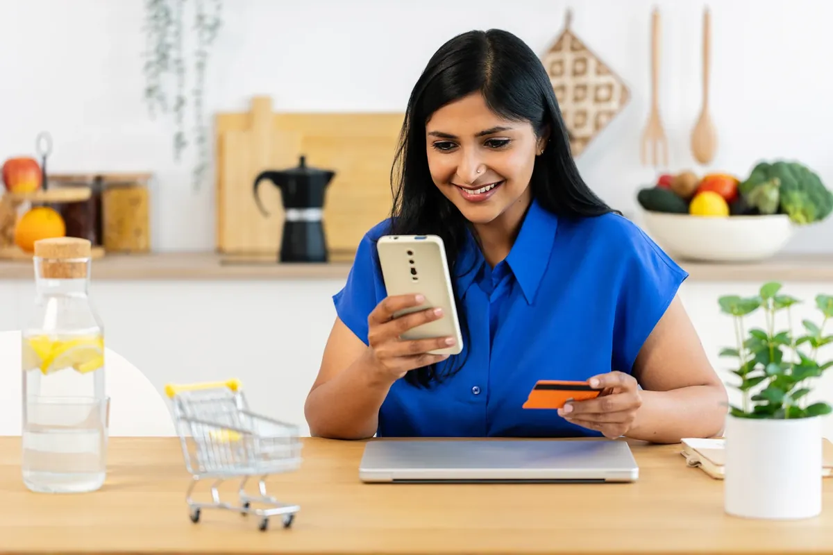 Smiling woman in a blue shirt holds her phone and a bank card at a kitchen table, placing an online order