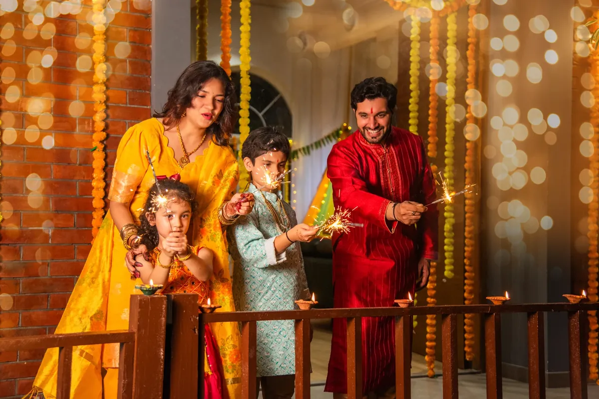 A family in festive clothes holds sparklers by a railing lined with diyas, with marigold garlands and fairy lights