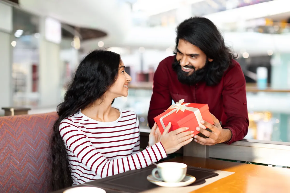 A man giving a red wrapped gift box to a smiling woman in a mall cafe