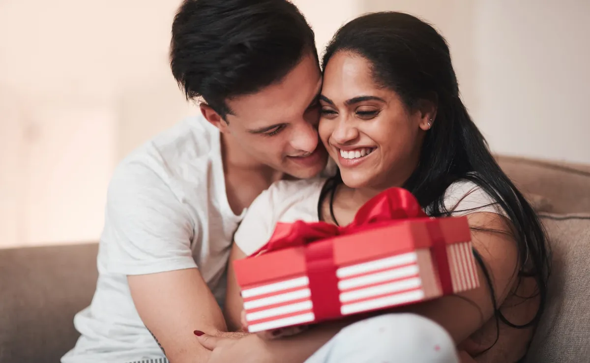 A smiling woman holding a red and white striped gift box with a red ribbon while a man embraces her from behind on a sofa
