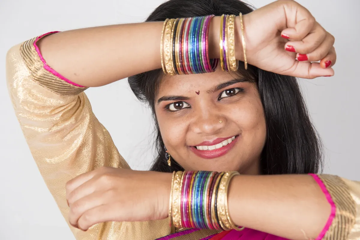 A smiling woman in a gold and pink top holding both wrists up to show stacks of gold and colourful glass bangles