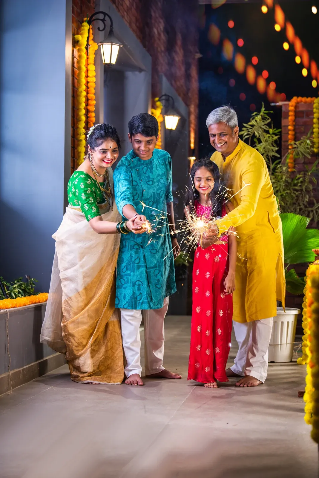 A family of four in festive Indian clothes holding lit sparklers on a decorated, marigold-lined walkway at night