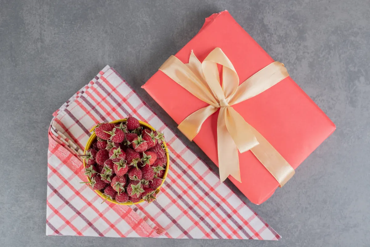 A red wrapped gift with a cream satin ribbon next to a yellow bowl of fresh raspberries on a folded checked cloth