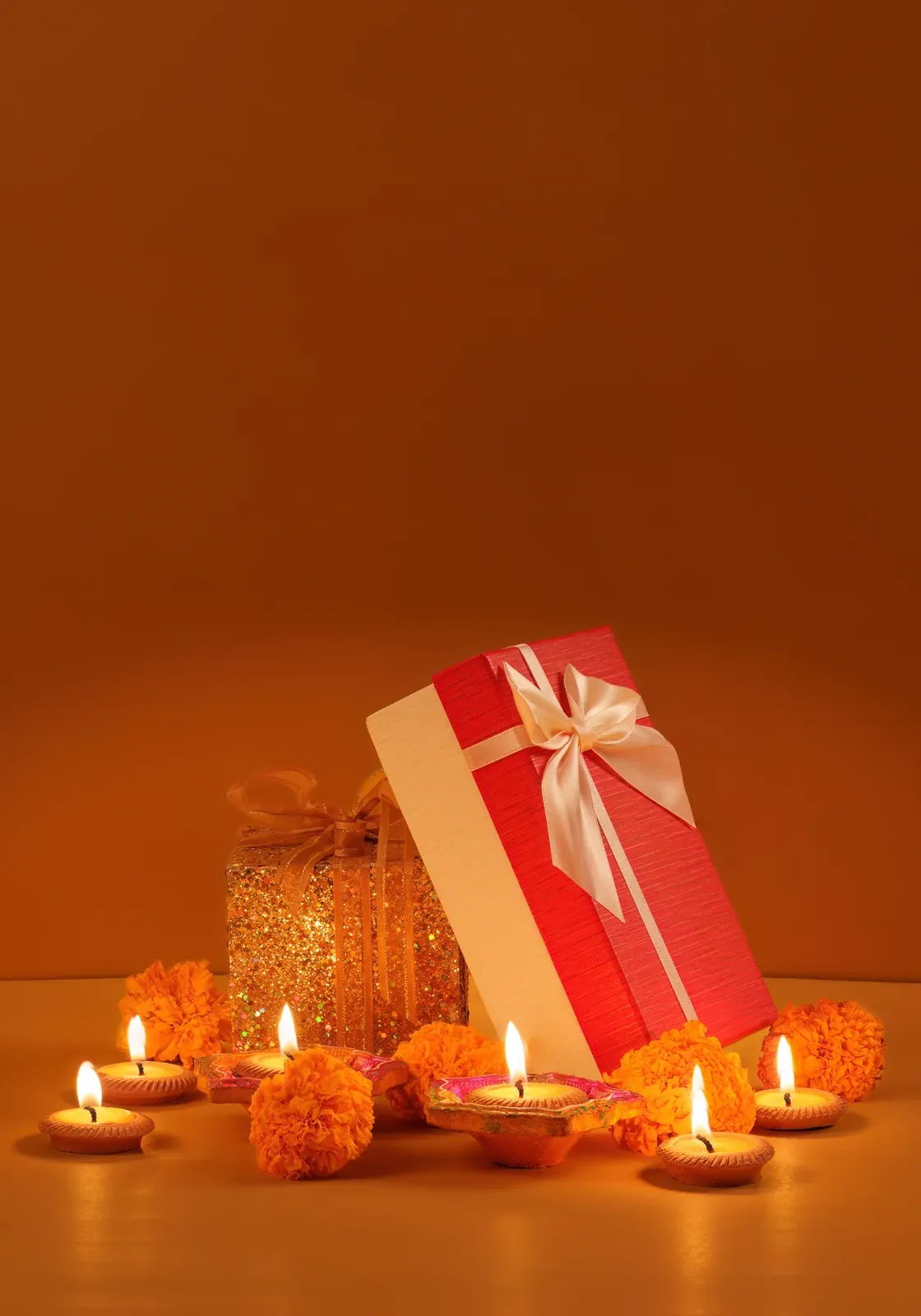 A red gift box with a cream ribbon beside a gold glitter-wrapped box, with lit clay diyas and orange marigolds on an orange background