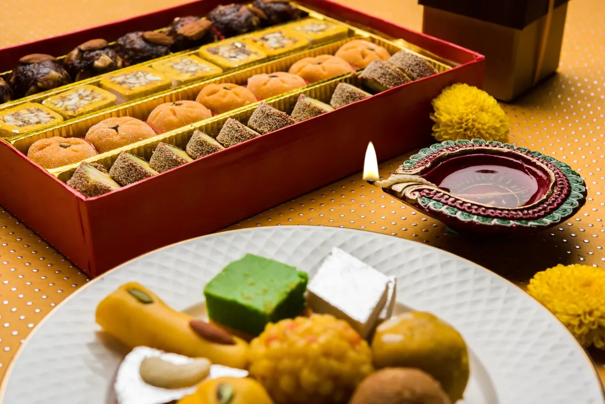 A red box of assorted Indian sweets and a plate of ladoo, barfi and silver-leaf mithai beside a lit diya and marigolds