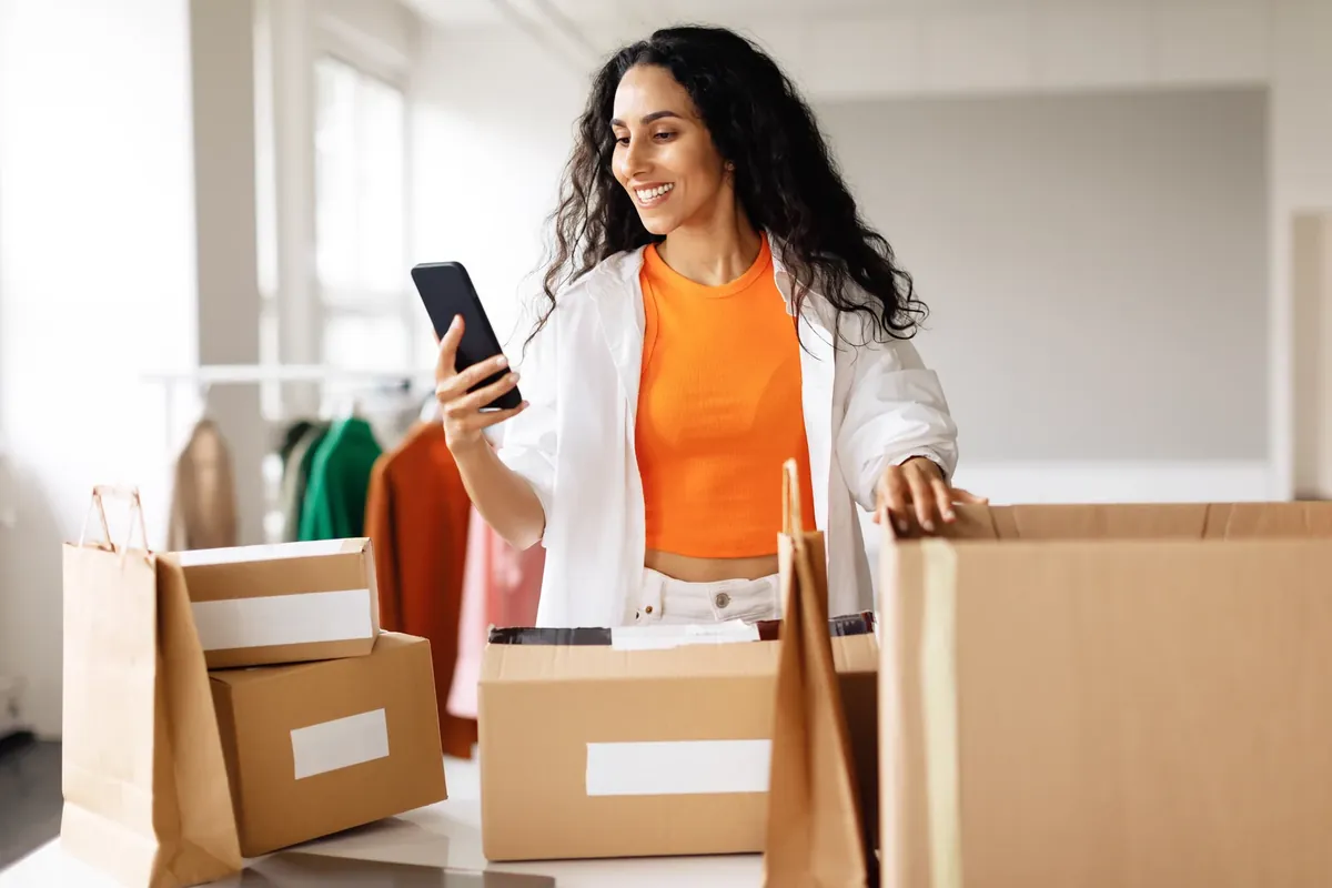 A smiling woman with long curly hair looks at her phone while packing cardboard boxes and paper bags in a bright room