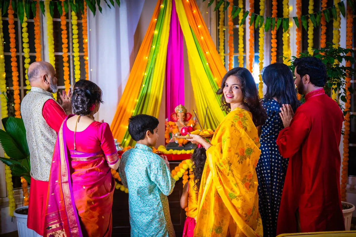 A family standing before a decorated Ganesha idol in a room hung with marigold garlands and draped orange, green and pink cloth with string lights, as a woman holds a puja thali