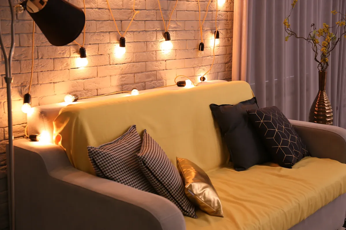 A sofa with a yellow cover and cushions in front of a white brick wall, with a string of bulbs hanging from the wall