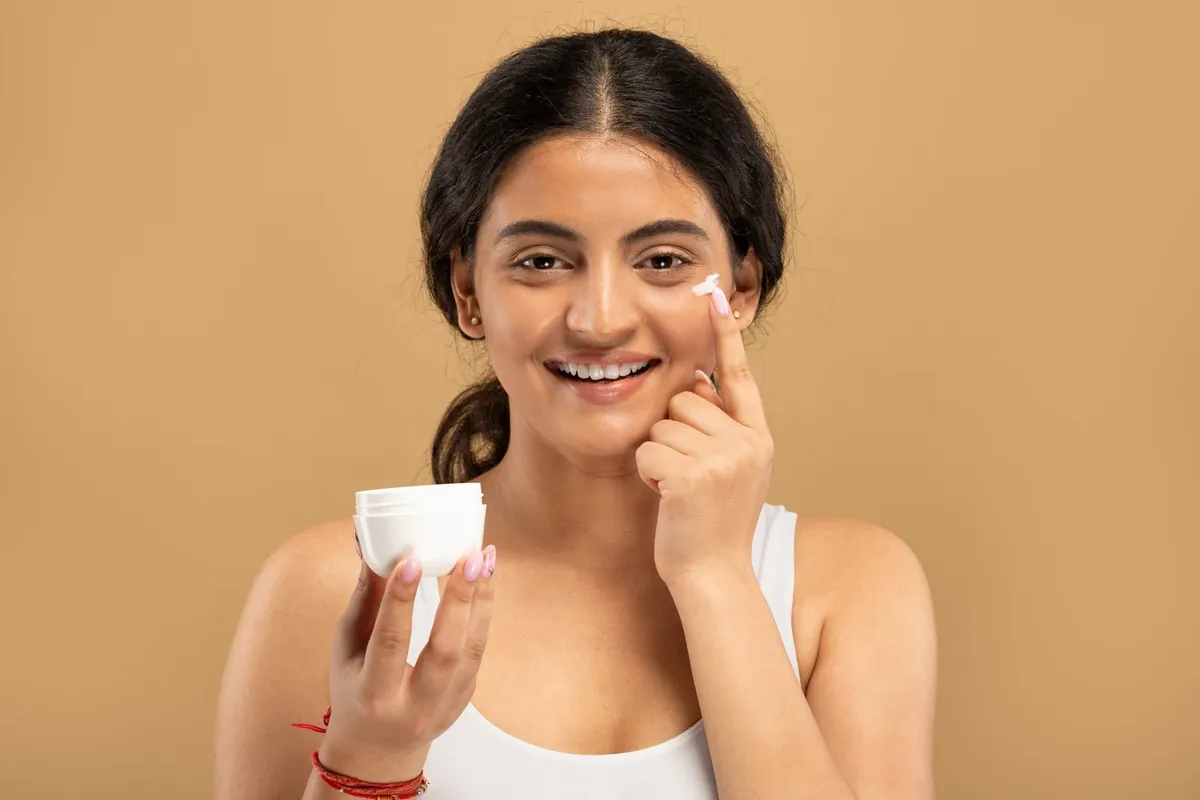 A smiling woman holding a white tub of cream and dabbing a little on her cheek against a warm tan background