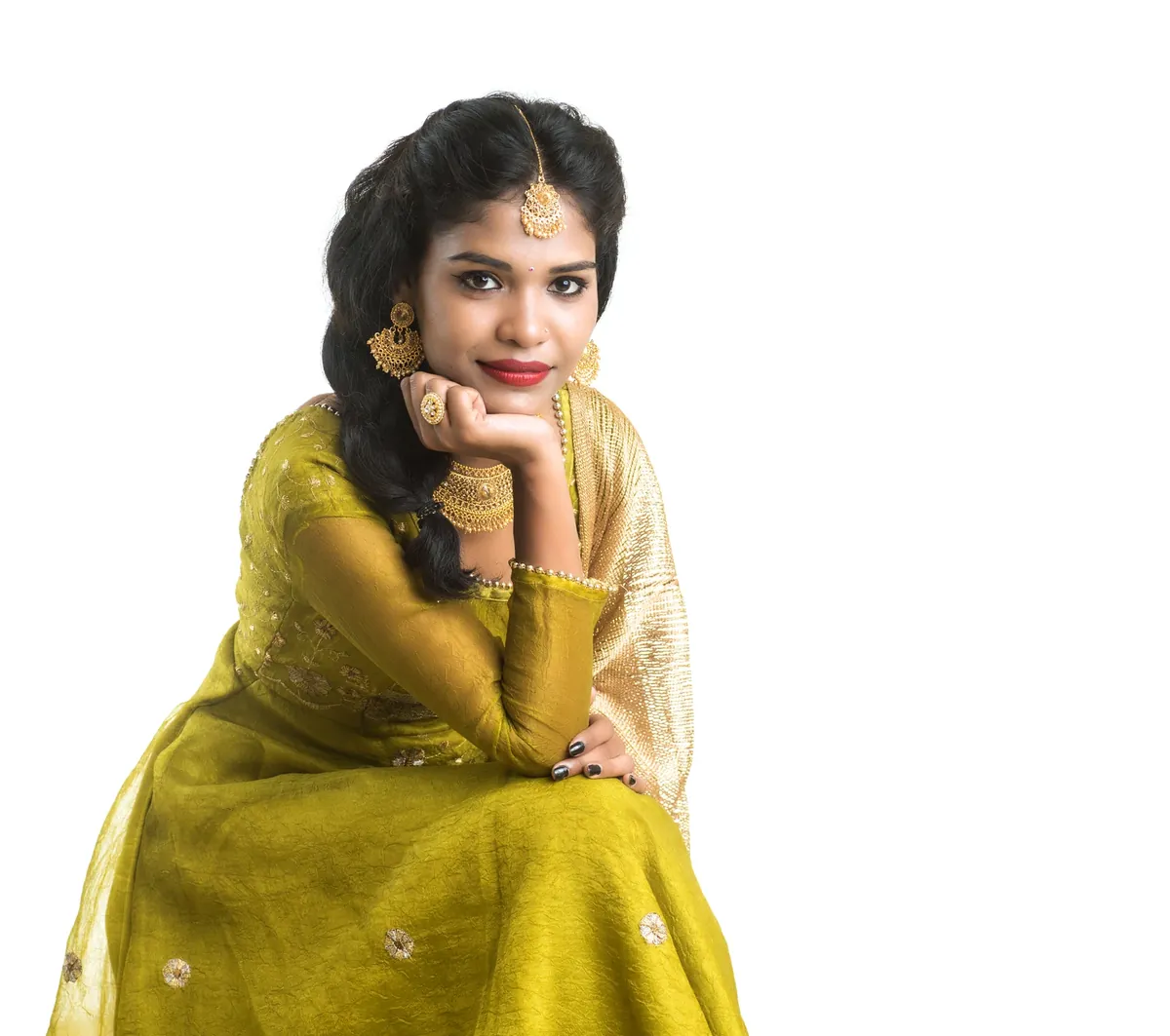 A smiling woman in an olive green outfit with a sheer, gold-bordered dupatta, a maang tikka and gold earrings, resting her chin on her hand