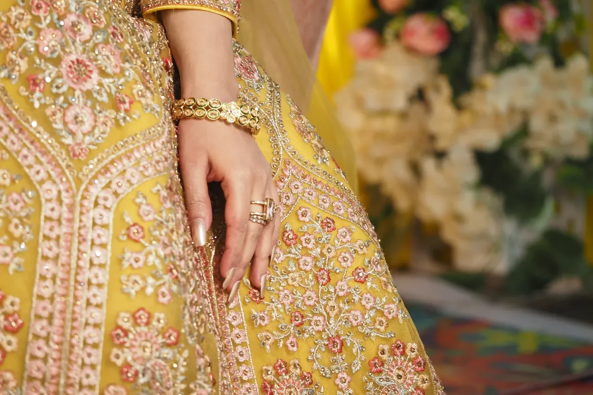 Close-up of a yellow lehenga with fine pink and gold floral embroidery, with a hand wearing a gold bracelet and rings