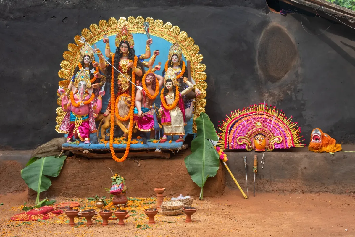 Durga idol with other deities and a lion, surrounded by marigold garlands, clay lamps and a colourful mask on a mud platform