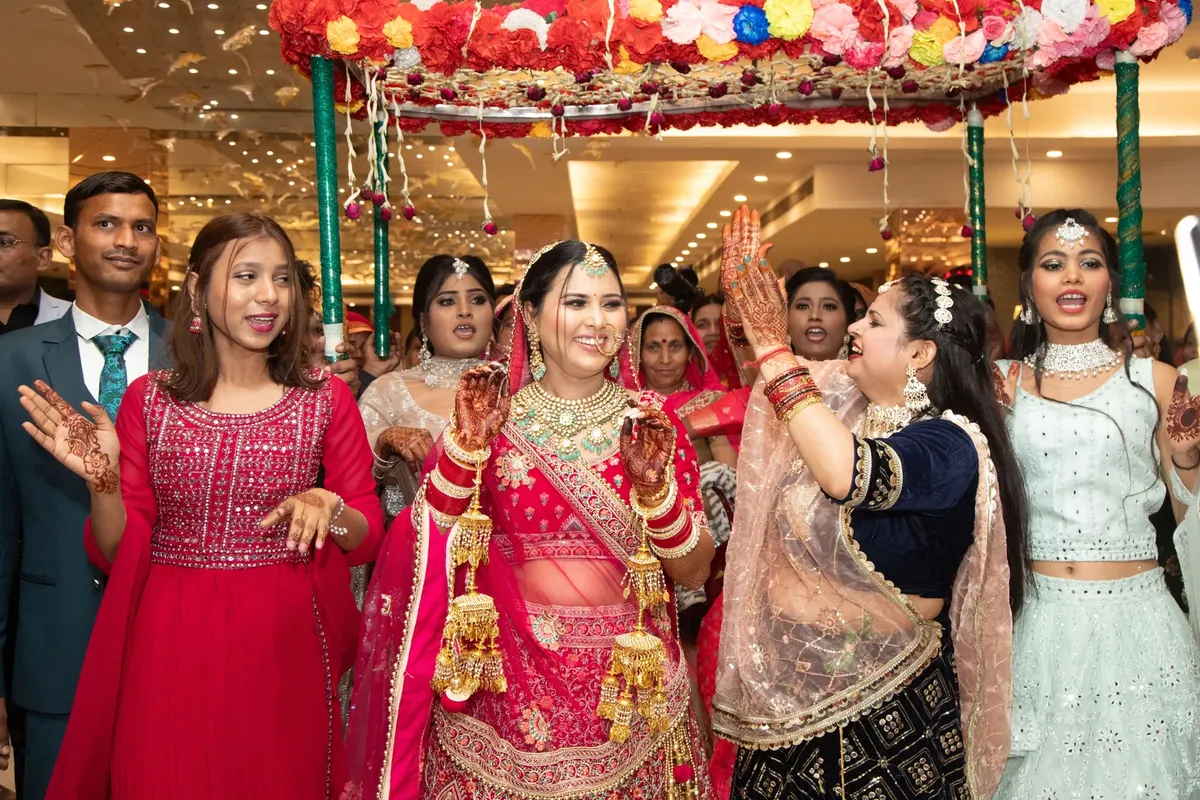 A bride in a red lehenga with gold jewellery dances with relatives and guests under a flower canopy at an Indian wedding