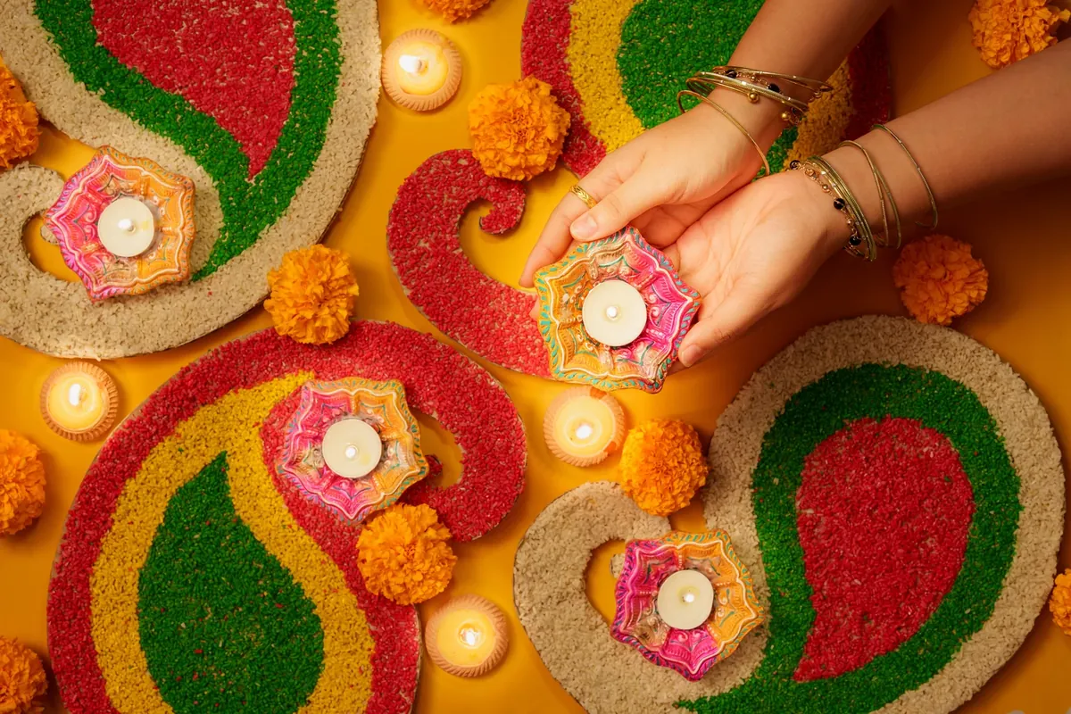 Hands placing a lit diya on a colourful rangoli of green, red and yellow paisley shapes with marigolds
