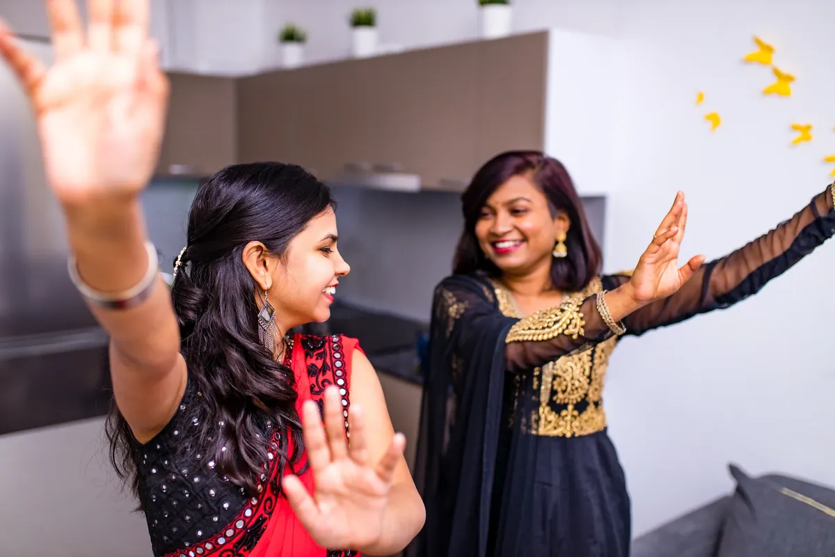 Two women in black and gold festive outfits laugh and dance, one with jhumkas and hair pinned half up