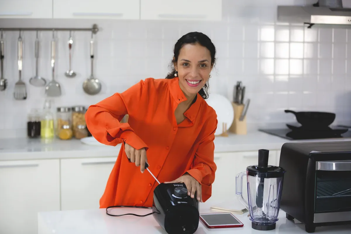 A smiling woman in an orange shirt in a bright white kitchen, assembling a blender on the counter next to a toaster oven