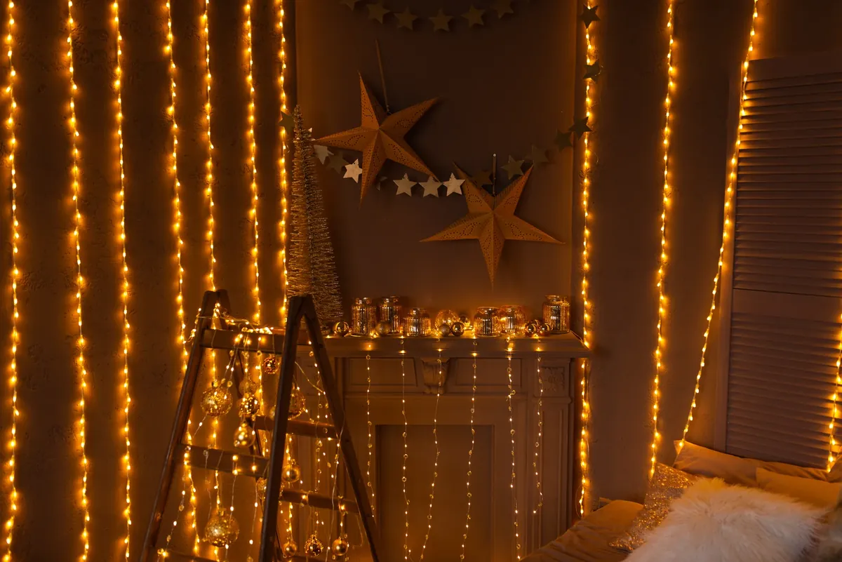 Warm fairy lights hanging in strings around a fireplace mantel decorated with paper stars, small gold baubles and a tiny tree