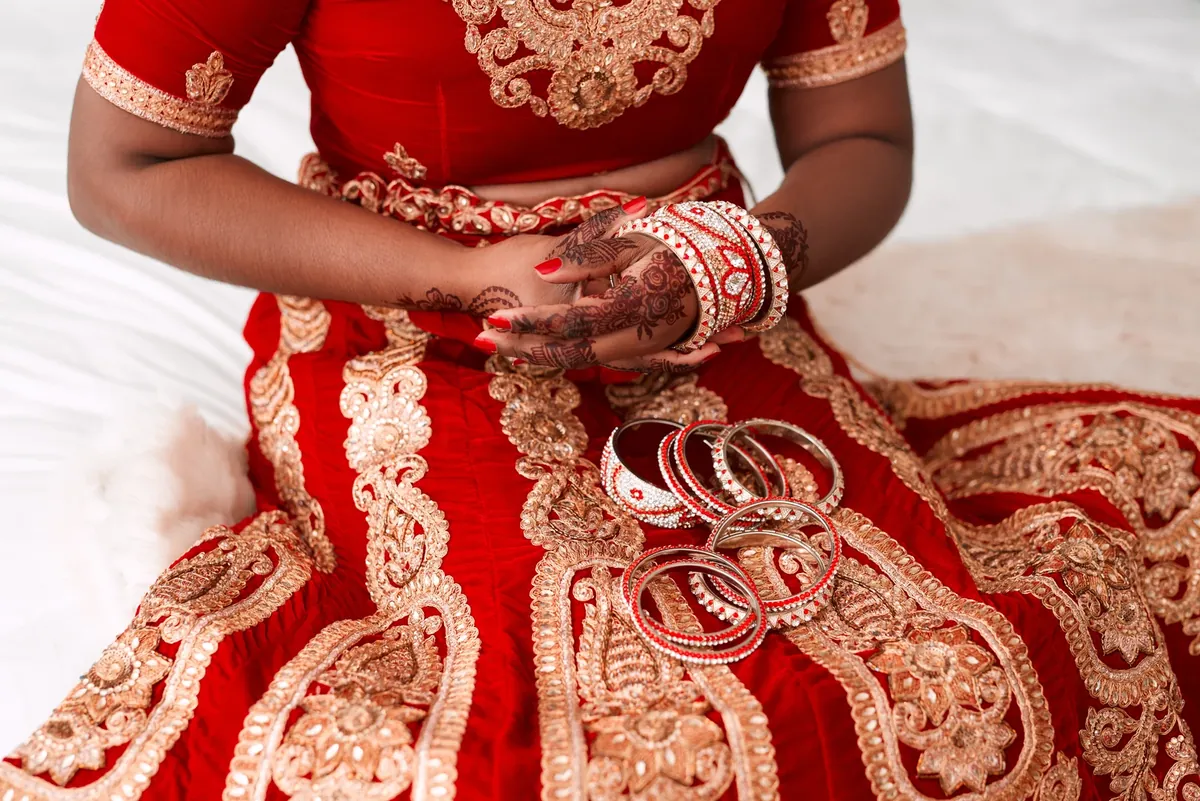 A woman in a red and gold lehenga with henna on her hands, wearing red and white bangles, with more bangles resting on her lap