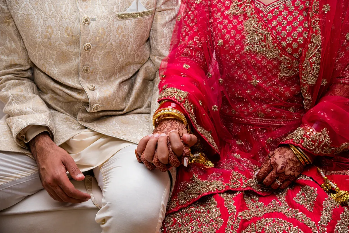 A couple seated side by side holding hands at a wedding ceremony, the woman in a deep pink embroidered outfit with henna on her hands and a gold bangle on her wrist