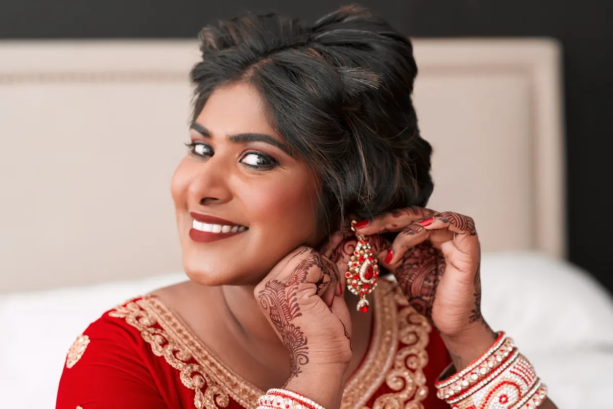 A smiling woman in a red outfit with henna on her hands fastening an ornate earring, with red and white bangles on her wrist