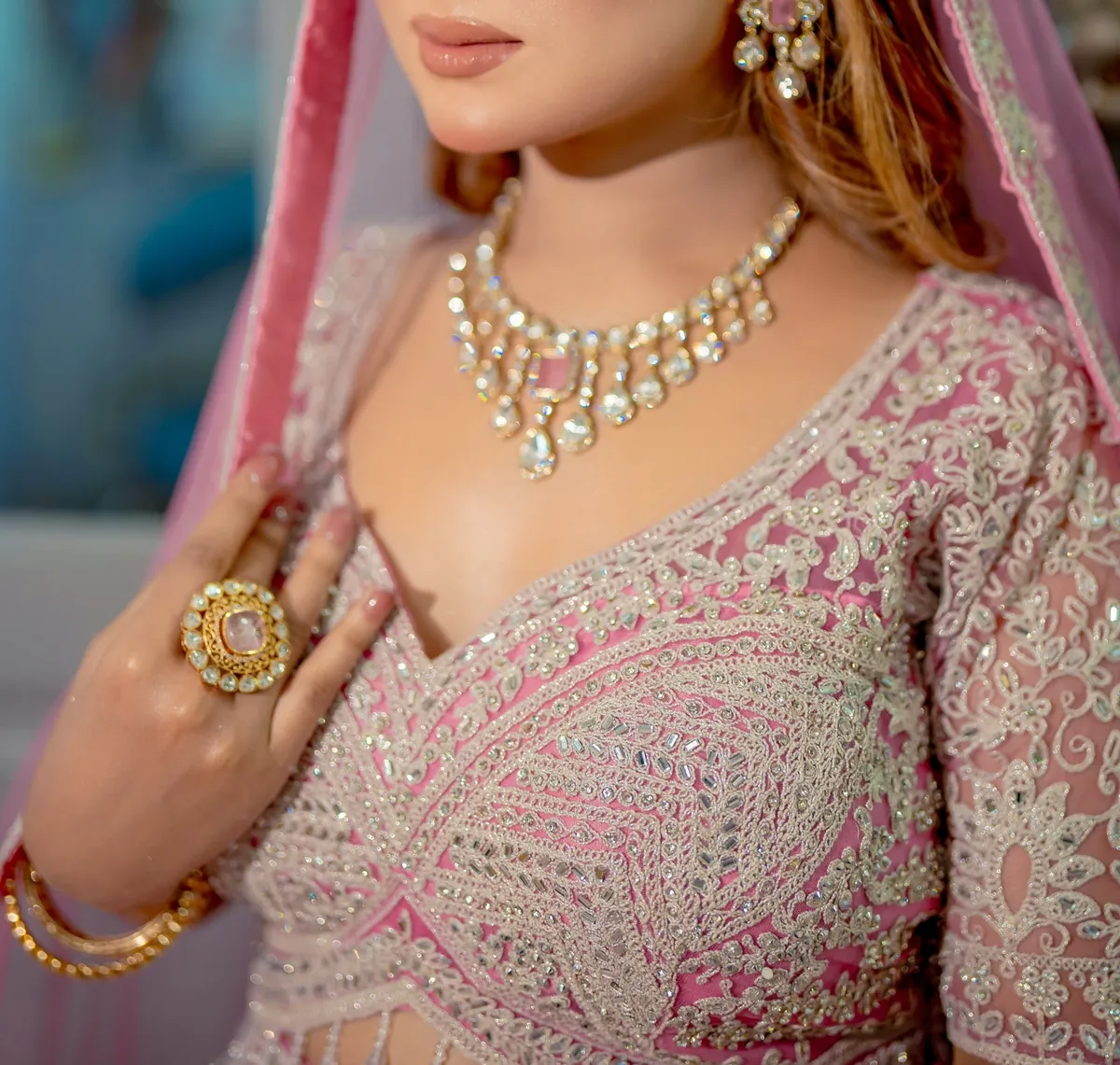 A woman in a pink embroidered outfit wearing a layered stone-set necklace, a cocktail ring and a bangle, photographed from the lips down
