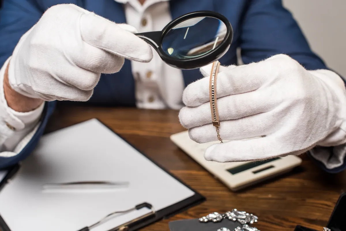 Jeweller in white gloves examining a gold chain bracelet through a magnifying glass