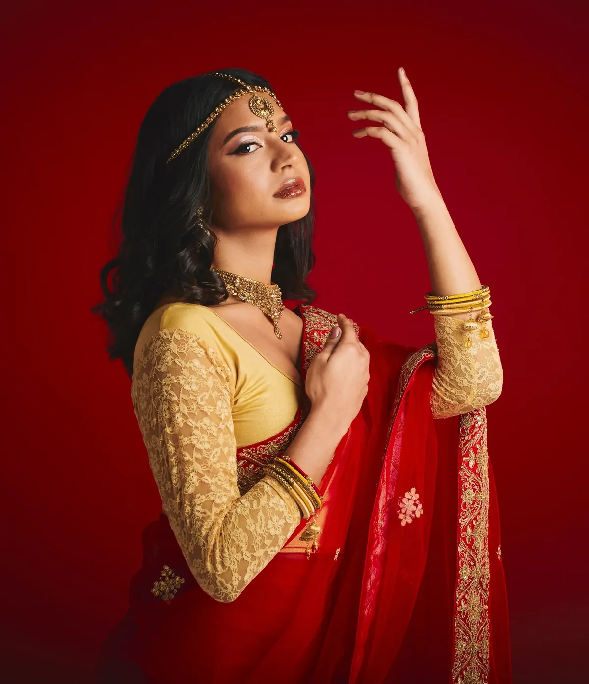 Woman in a red saree with a gold-embroidered border and a gold lace blouse, wearing a maang tikka, against a red wall