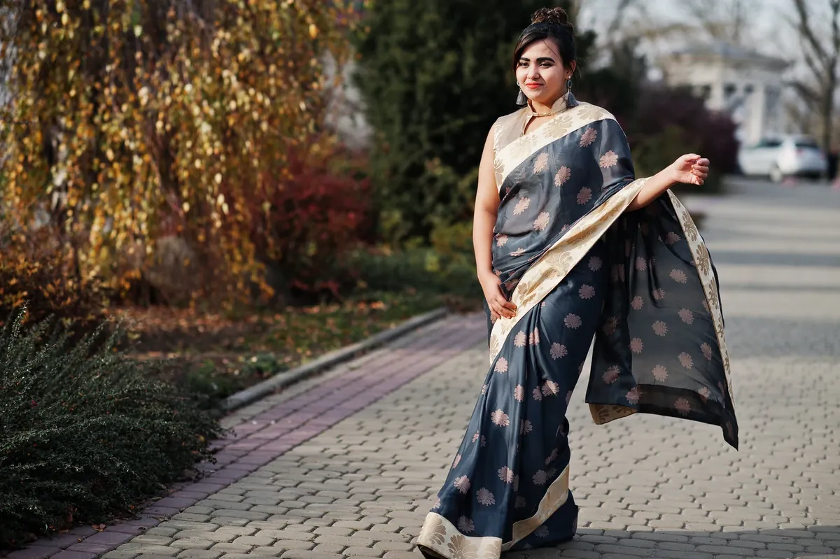 Woman in a grey saree with small gold motifs and a cream border, standing on a garden path with the pallu flowing