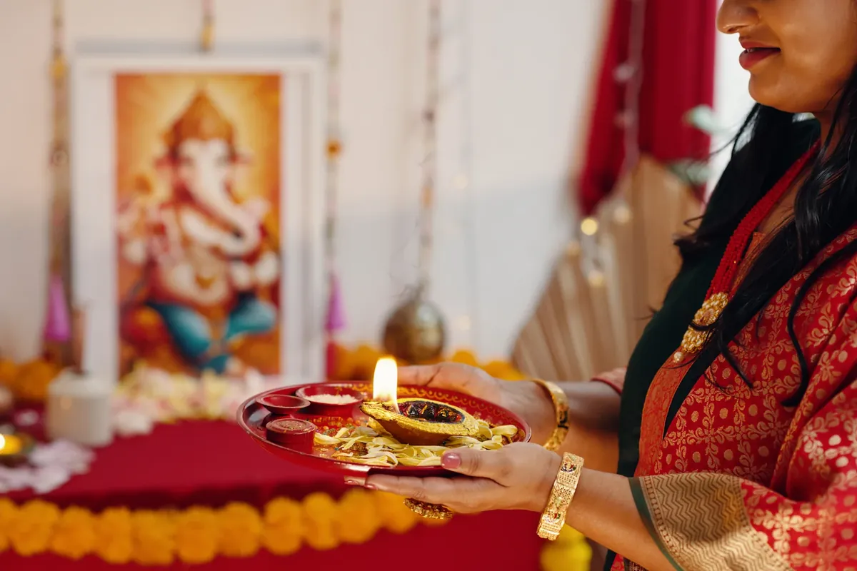 A woman in a red and gold sari holds a lit oil lamp on a small plate in front of a framed picture of Ganesha and a red cloth-covered table