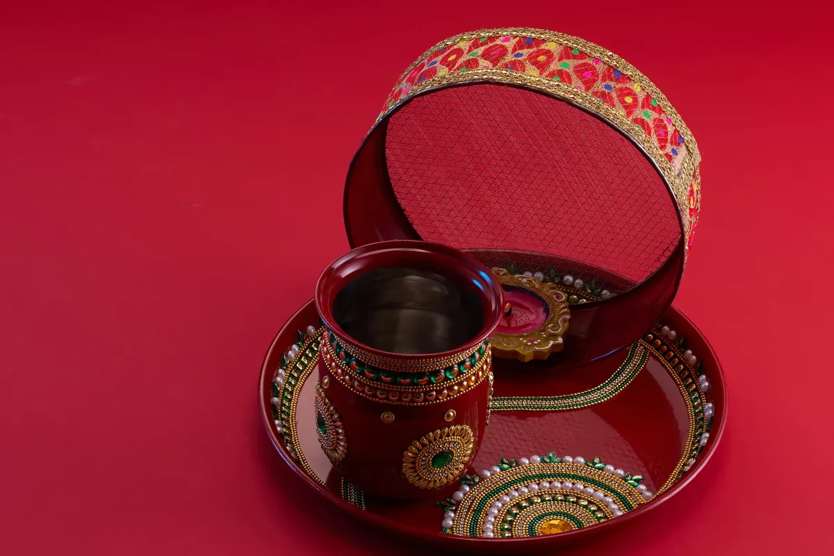 A red decorated puja thali with a painted karwa pot and an embroidered sieve against a red background